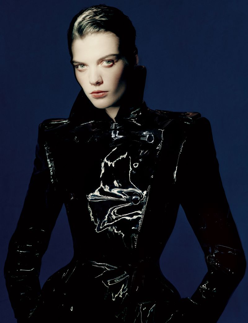 A woman in a shiny black coat stands against a deep blue background in a fashion-style studio shot with a strong light source.