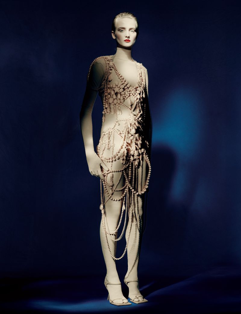 A woman in a dress made of pearls stands in a full shot against a blue background with rim lighting creating a dramatic fashion image.