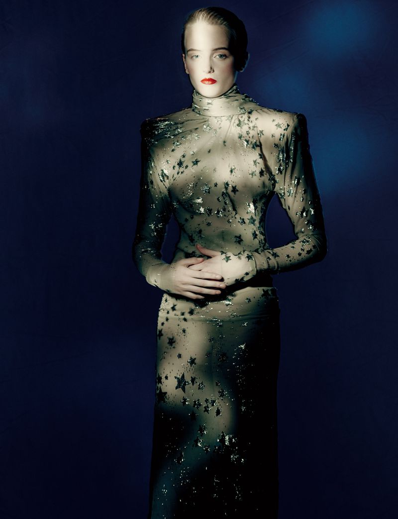 Adult fashion model in sheer star-patterned turtleneck dress stands with hands clasped in a studio shot against a dark blue backdrop.