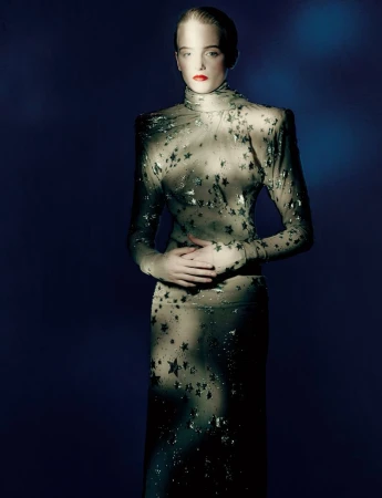 Adult fashion model in sheer star-patterned turtleneck dress stands with hands clasped in a studio shot against a dark blue backdrop.