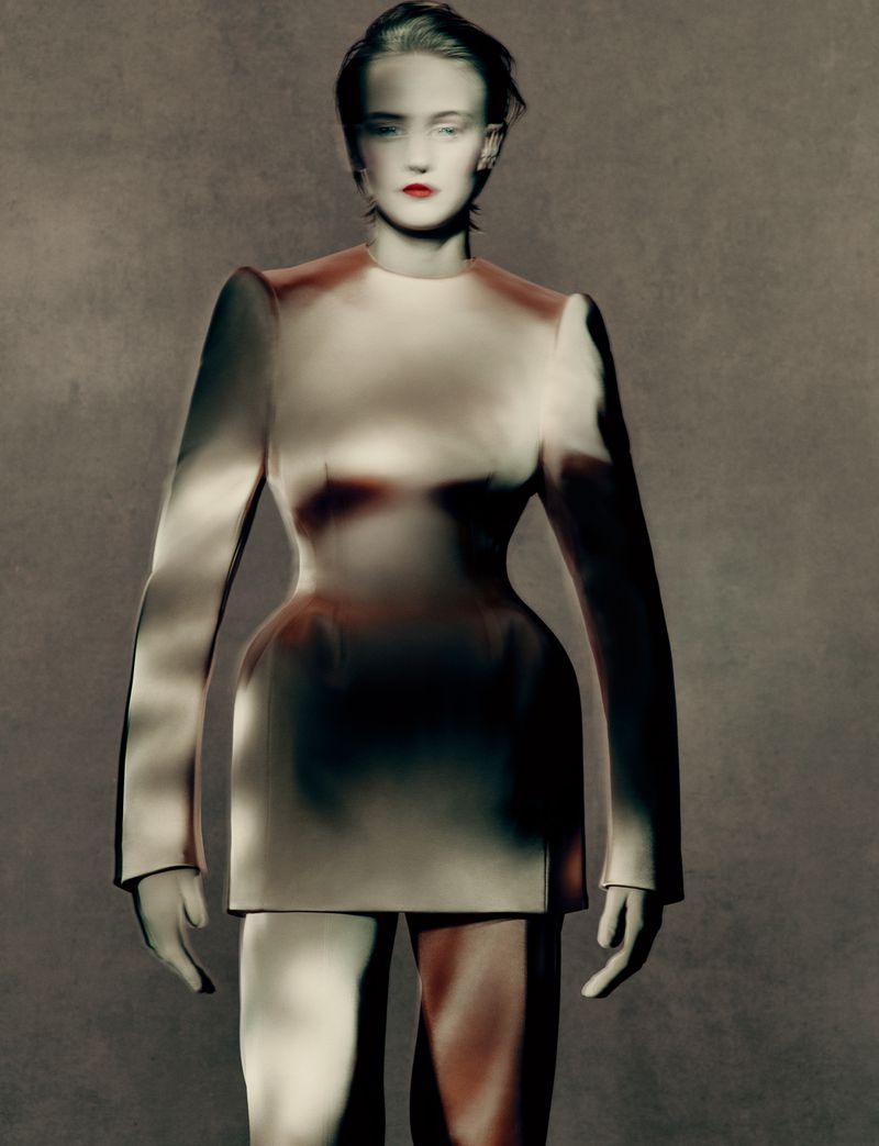 Studio shot of a pale-skinned adult female in a tailored white dress with long sleeves and brown shading, standing against a mottled grey backdrop.