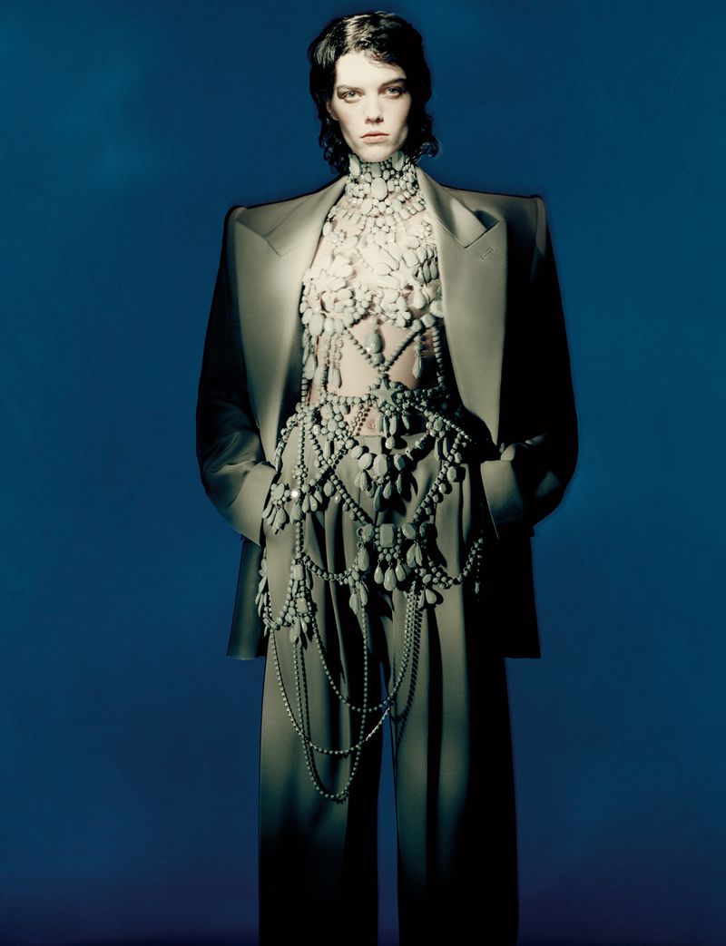 A woman poses in a grey pantsuit with ornate pearl detailing, standing against a dark blue backdrop, with a direct gaze at the camera.