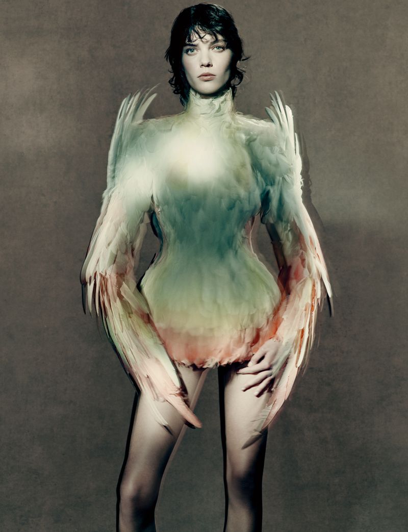 A female model poses in a feathered dress against a grey background, looking directly at the camera in a studio setting.