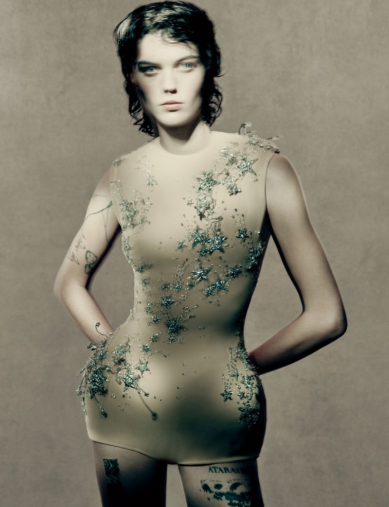 Fashion portrait of an adult female model with tattoos, in a beige embellished bodysuit, standing in a studio with a neutral background.