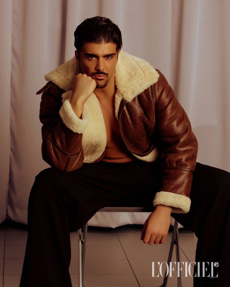 Portrait of a bearded adult male wearing a brown leather aviator jacket with cream shearling, seated on a metal folding chair in studio.