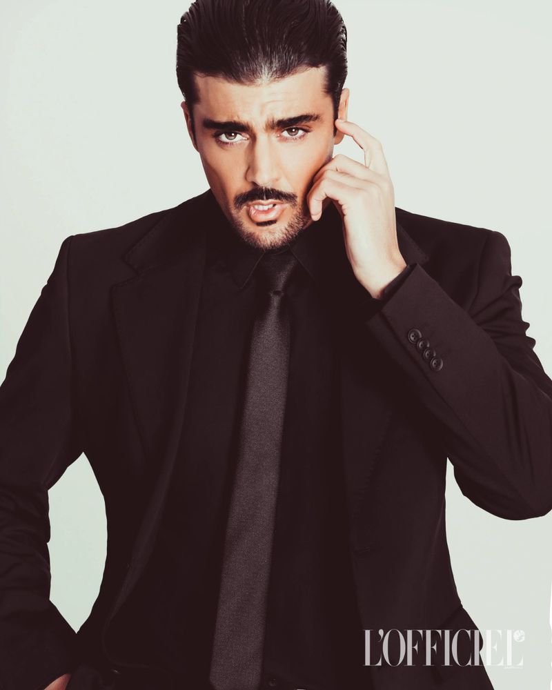 Man with dark hair and mustache wearing a black suit jacket and tie against a plain white background