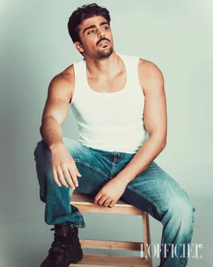 Man wearing a white tank top and denim jeans seated on a wooden stool against a light grey seamless background