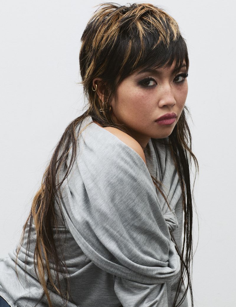 A young adult woman with dark and blonde hair and neutral expression poses in a gray top against a plain white background in a studio setting.