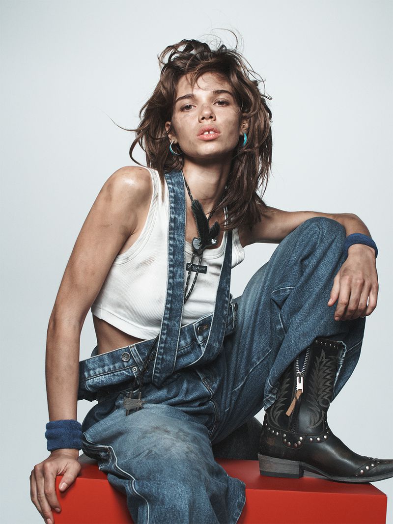 A woman with fair skin, overalls, and black boots sits on a red block against a white backdrop in a fashion-style portrait.