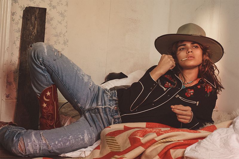 Adult female wearing cowboy hat and western shirt poses on bed with red cowboy boots and ripped jeans in an indoor setting with white walls.