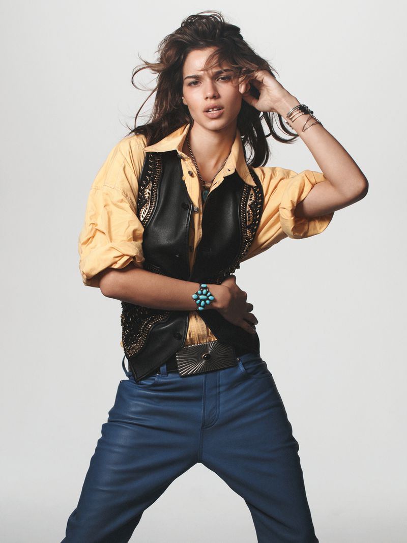 Fashion model in a yellow button-down shirt and black leather vest poses in front of a white background. She wears blue leather pants.