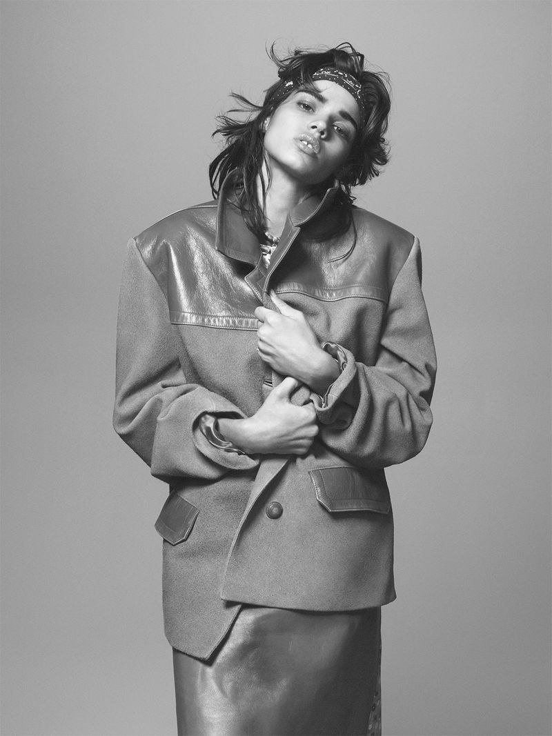 Black and white portrait of a woman wearing an oversized blazer, skirt, and bandana, posing against a neutral gray background.