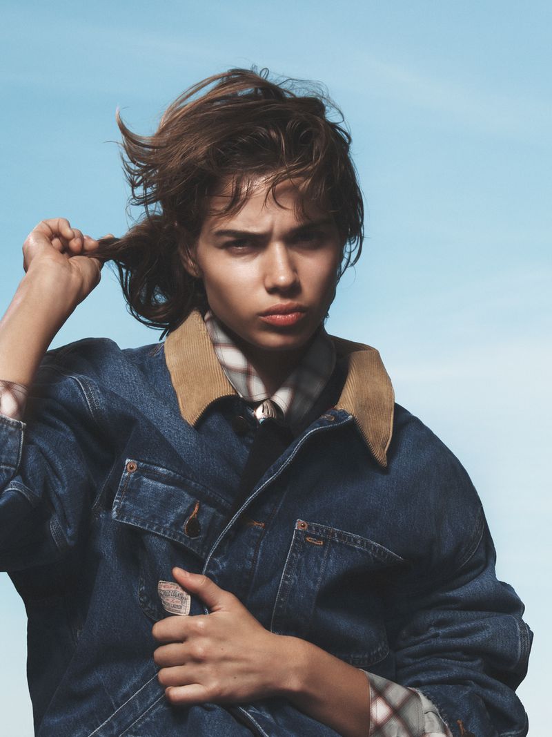 Portrait of a young woman with tousled brown hair, wearing a denim jacket, posing against a soft blue sky outdoors.
