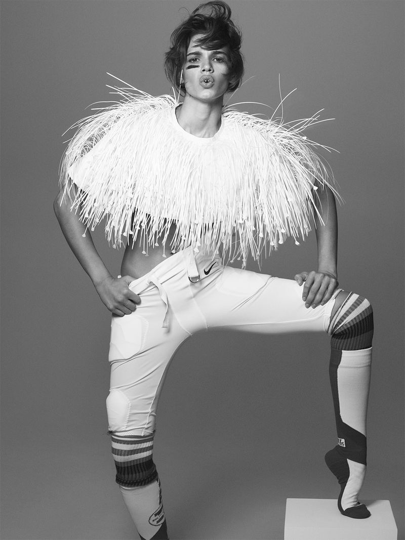 Fashion model poses in white athletic pants and shoulder pads against grey backdrop with dramatic hair and makeup in a studio setting.
