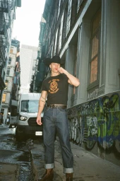 Man wearing a black cowboy hat and graphic t-shirt standing in a city alleyway with a parked van