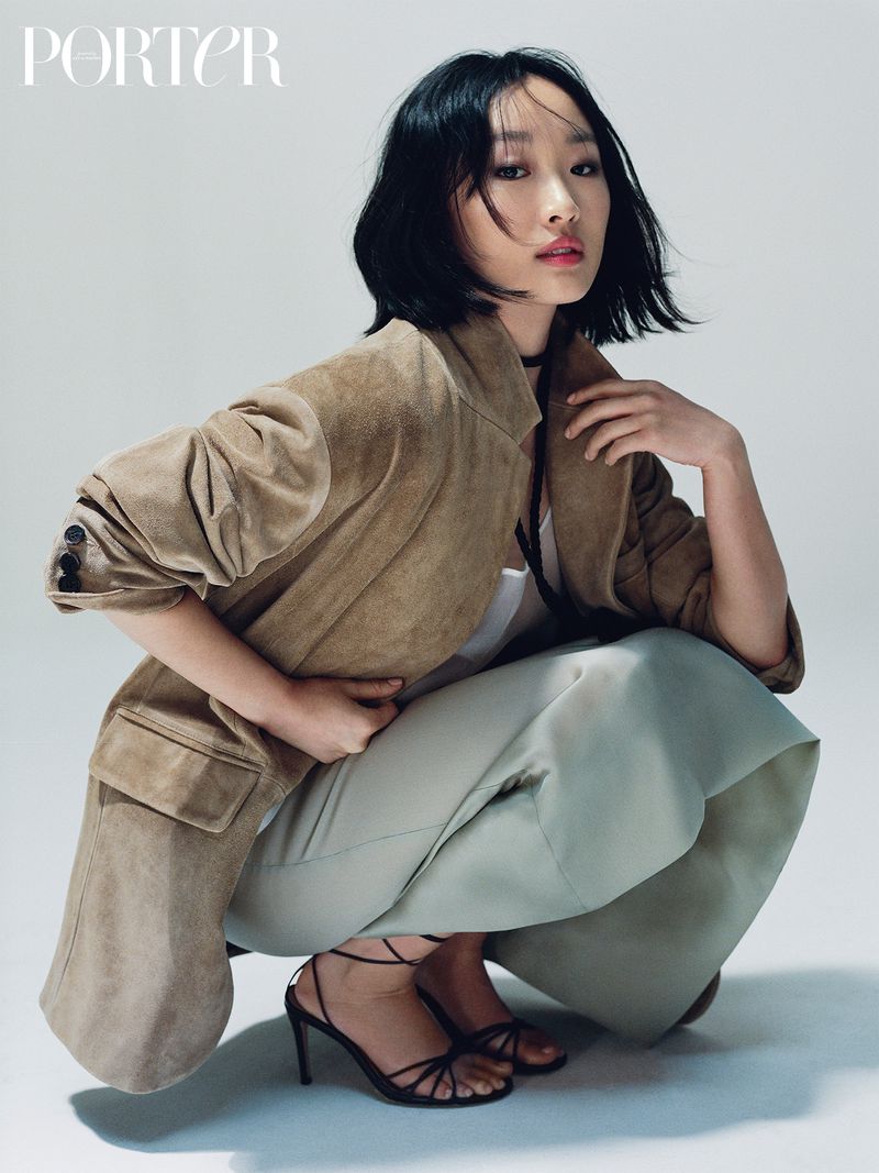 Elegant Asian woman in a tan jacket and light green pants poses in a studio wearing black heels for Porter magazine cover shoot.