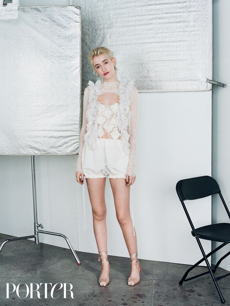 Woman in white lace top and shorts in a photo studio. The full shot features a silver background and black folding chair.