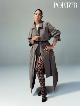 A woman poses in a gray trench coat and animal print tights in a studio setting with magazine branding in the upper corner.