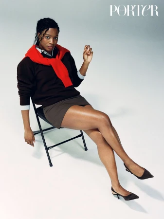 Alexandra Shipp seated on a folding chair wearing a dark sweater with a red sweater tied over her shoulders against a white background.