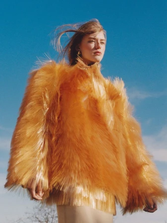 Woman in orange textured coat poses against a clear blue sky. Fashion shot with warm color grading. Outdoor fashion editorial.