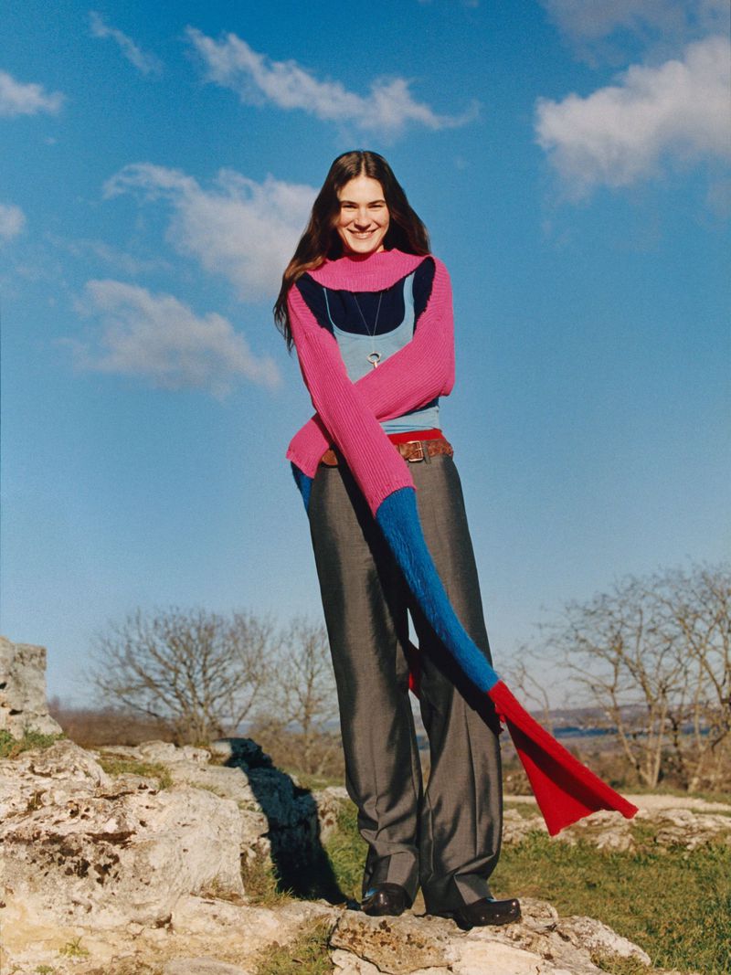A smiling young woman stands on a rocky hilltop wearing wide-legged pants and a colorful sleeve-like garment against a blue sky backdrop.