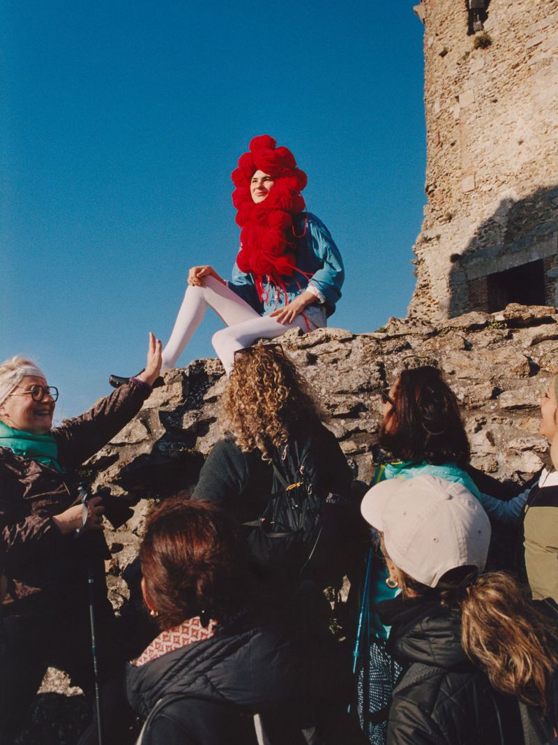 Woman with large red yarn headpiece sits on a rock wall in front of a blue sky, while a group of people stand below her.