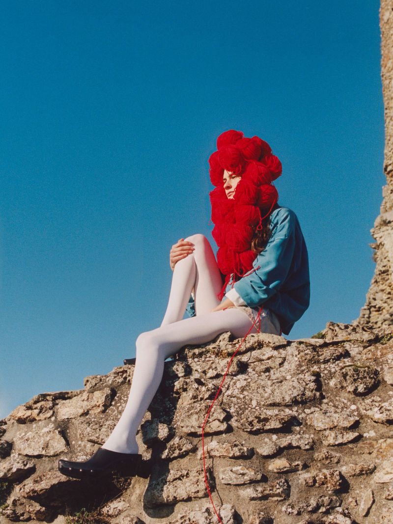 A woman in a red wool-like headdress sits on a stone wall against a clear blue sky, wearing a blue jacket and white tights. 