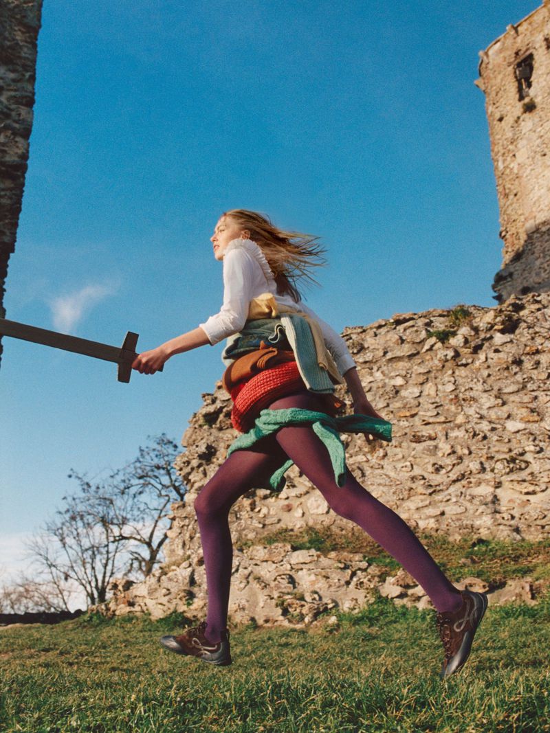 Woman in purple tights runs with sword near castle ruins under bright blue sky in outdoor fashion shot. Green grass in foreground.