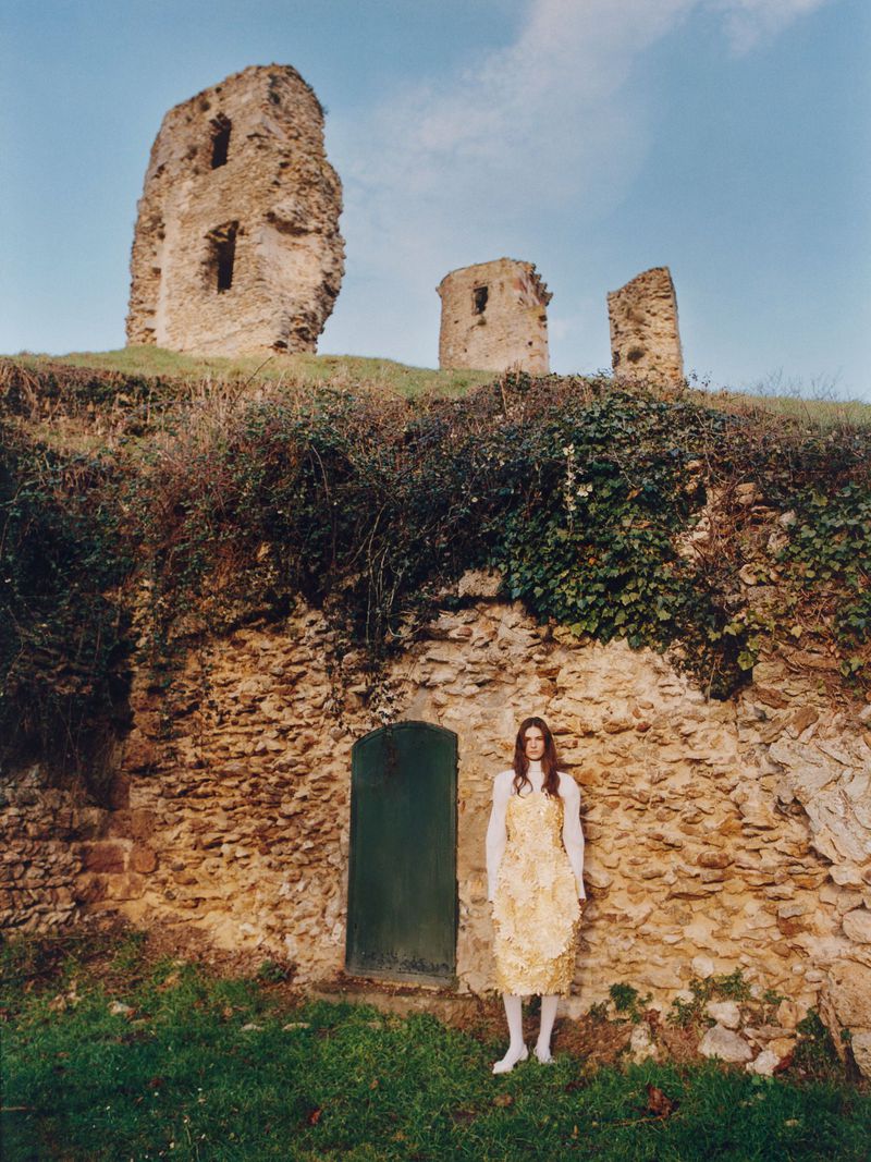 A woman in a yellow dress stands in front of a stone wall with a green door, with castle ruins visible on a grassy hill above.