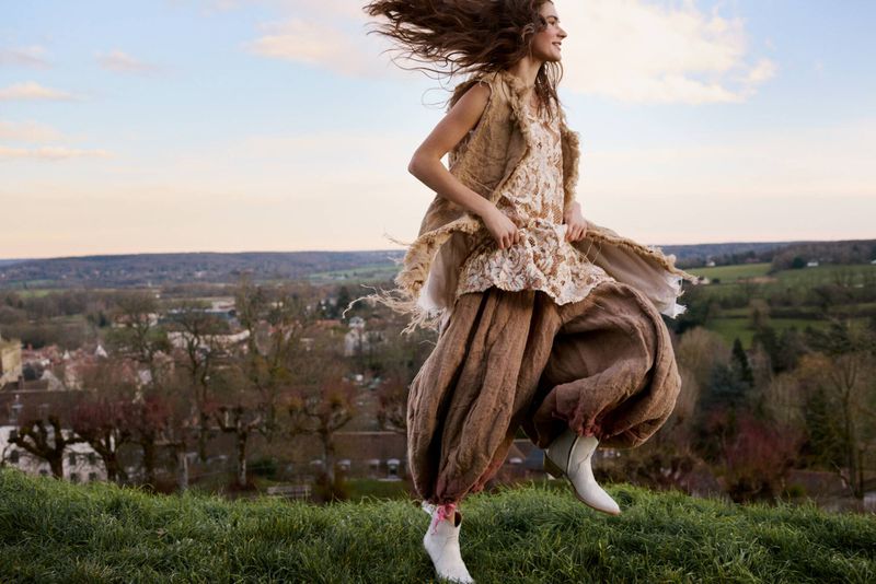 A woman jumps on a grassy hill wearing a flowing outfit consisting of a light-colored vest and top, and brown pants. A village is visible.