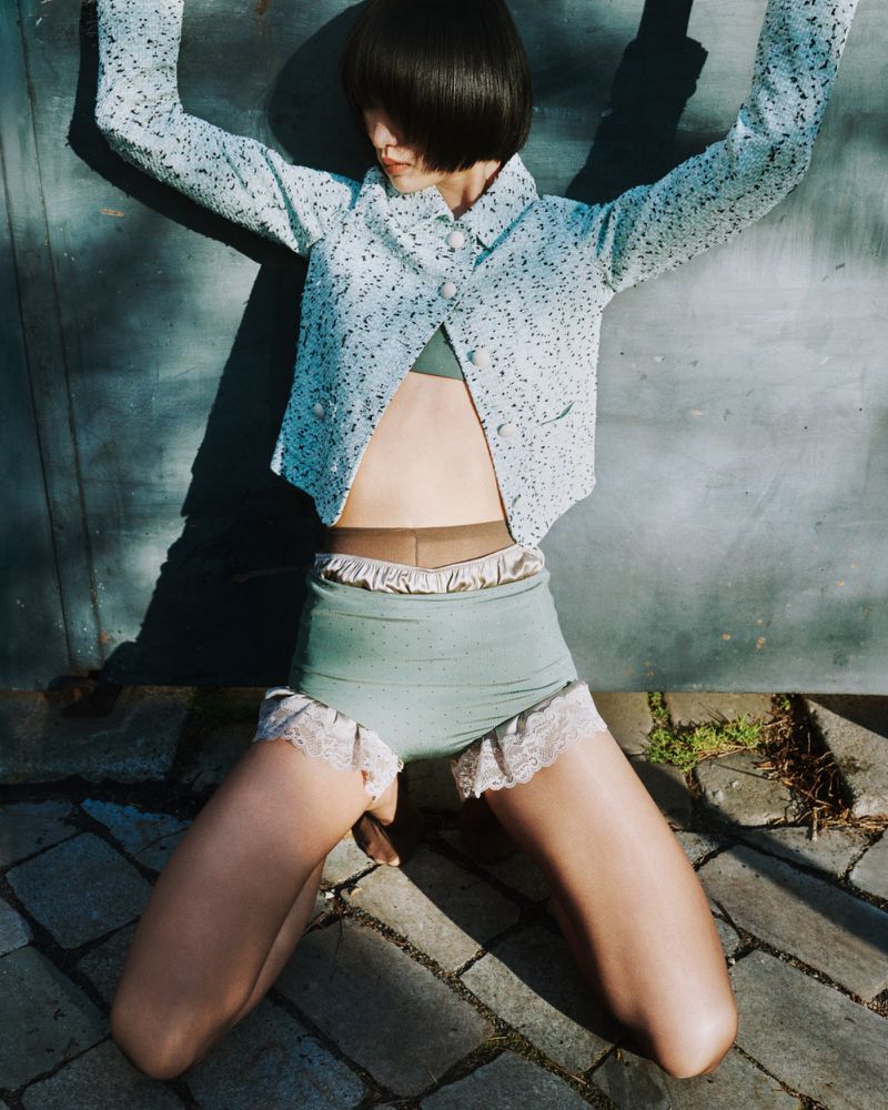 Young woman in high-waisted shorts and jacket kneels outside against a metal background with dappled sunlight and shadow.