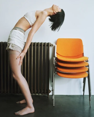 Young woman in neutral athleticwear arches her back while posing barefoot in front of a stack of orange chairs and a dark radiator.