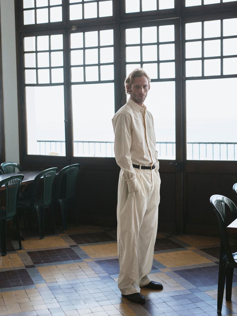 Full-length portrait of a fair-skinned adult male in a light-colored outfit, standing in an indoor space with large windows and tiled floor.