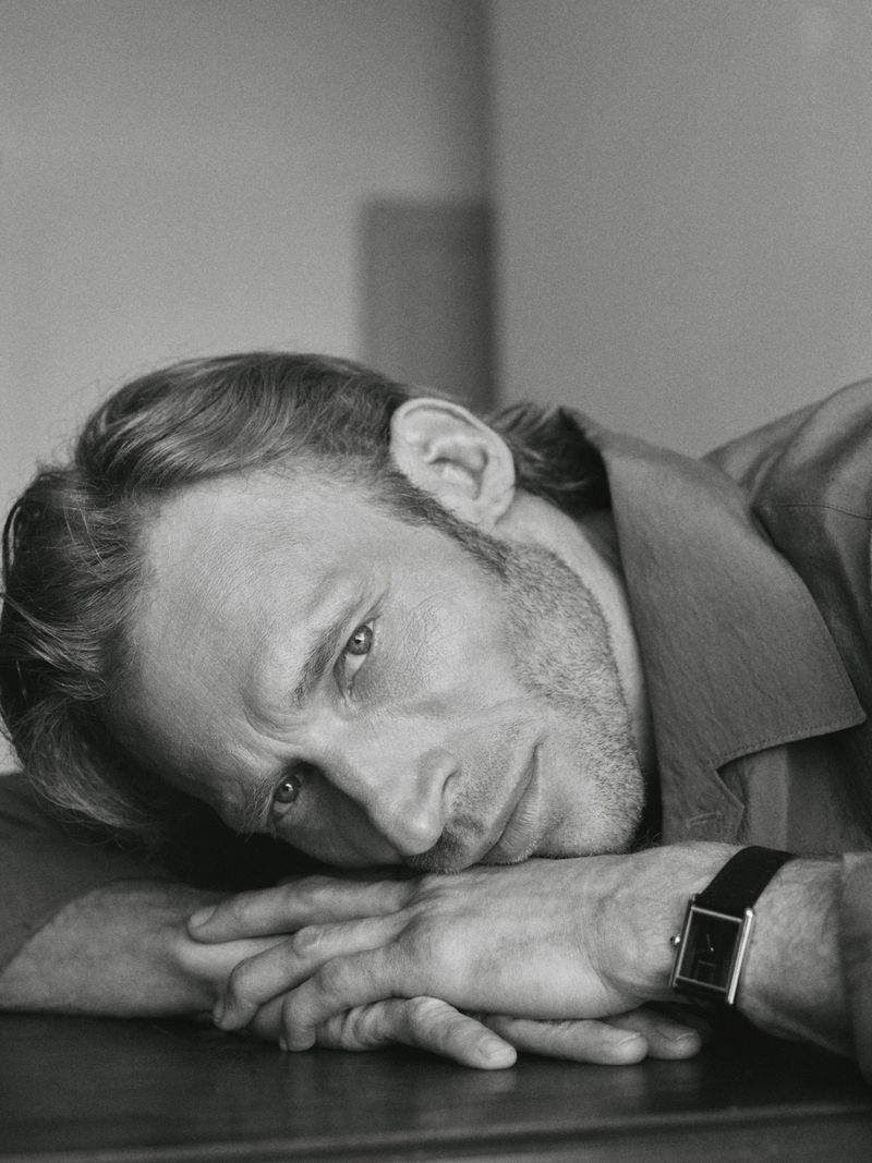 Monochrome portrait of a man with a beard, resting his head on his hands wearing a watch, captured in a close-up shot with soft lighting.