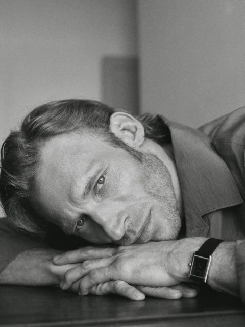 Monochrome portrait of a man with a beard, resting his head on his hands wearing a watch, captured in a close-up shot with soft lighting.