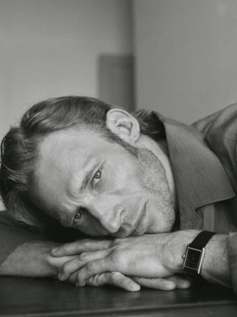 Monochrome portrait of a man with a beard, resting his head on his hands wearing a watch, captured in a close-up shot with soft lighting.