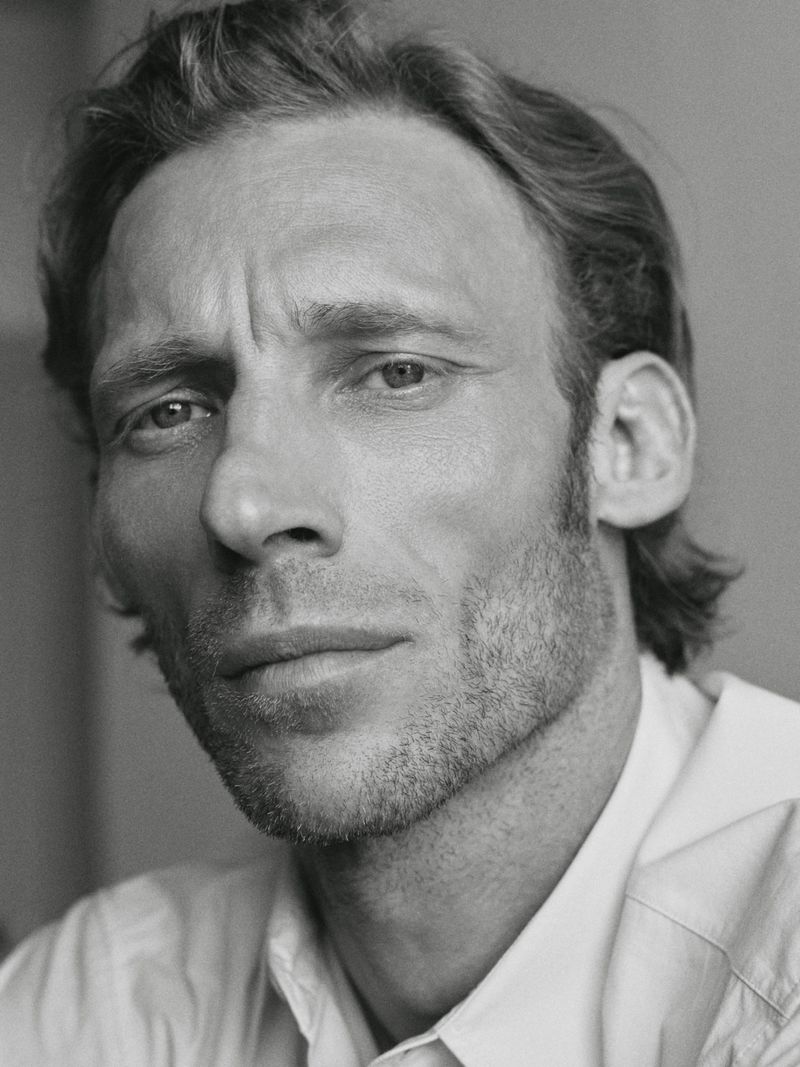 Close-up portrait of a man with wavy hair and stubble, wearing a light-colored button-up shirt, presented in monochrome.