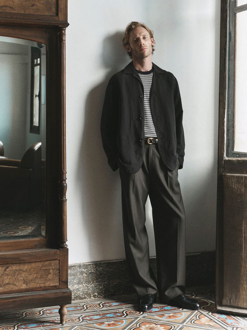 Adult man in a blazer leans against a white wall next to a cabinet in a relaxed, indoor pose, with a tiled floor and natural light.