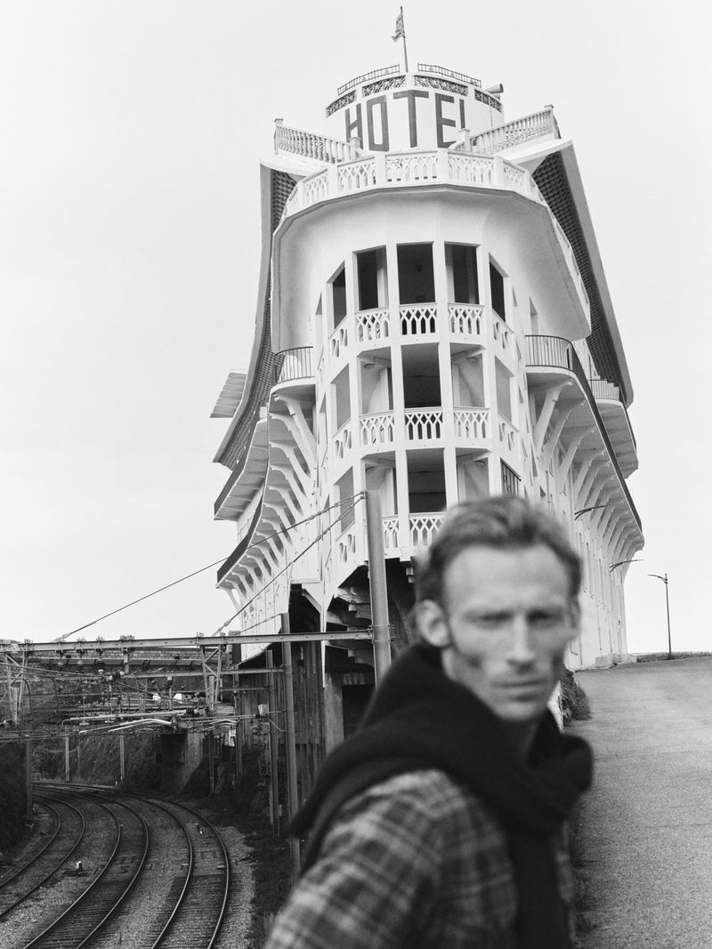 Monochrome portrait of a man with a black sweater, standing in front of a white hotel and railway tracks on a cloudy day.