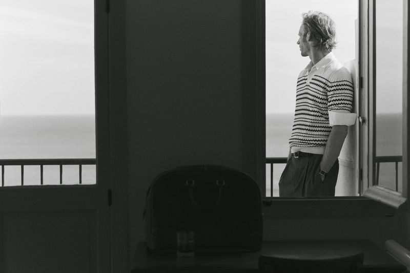 Monochromatic portrait of a man in a striped shirt leaning against a doorway, overlooking the ocean with a travel bag nearby.