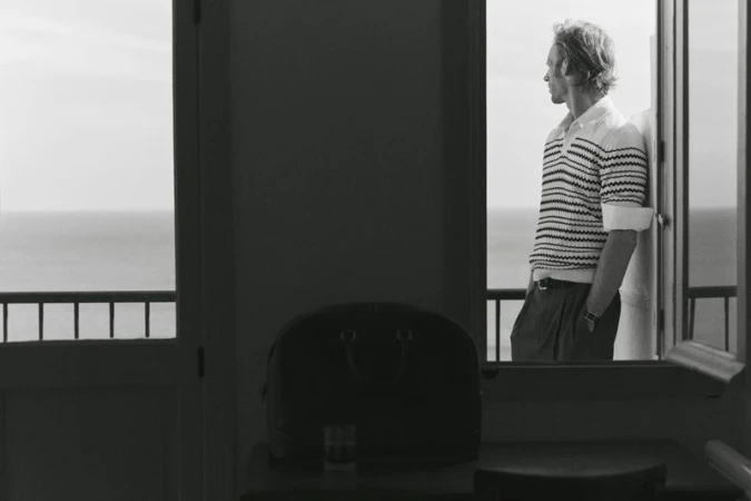 Monochromatic portrait of a man in a striped shirt leaning against a doorway, overlooking the ocean with a travel bag nearby.