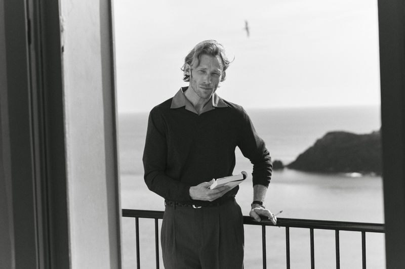 Monochrome portrait of a fair-skinned adult man with blond hair standing on a balcony, holding a notebook, with a coastal view in the background.