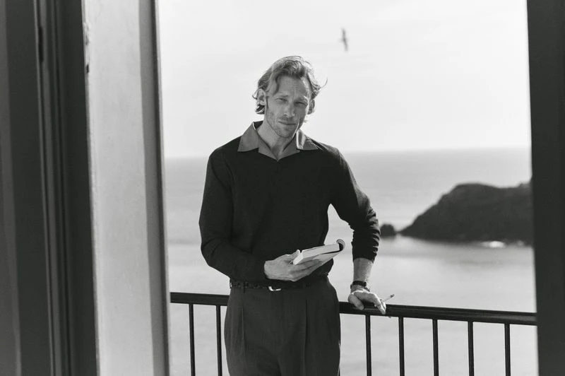 Monochrome portrait of a fair-skinned adult man with blond hair standing on a balcony, holding a notebook, with a coastal view in the background.