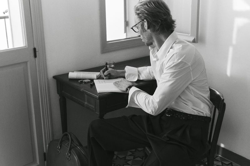 Man writing at a desk in a room with a doorway and framed picture; wearing glasses, shirt, and pants. Black and white photography.