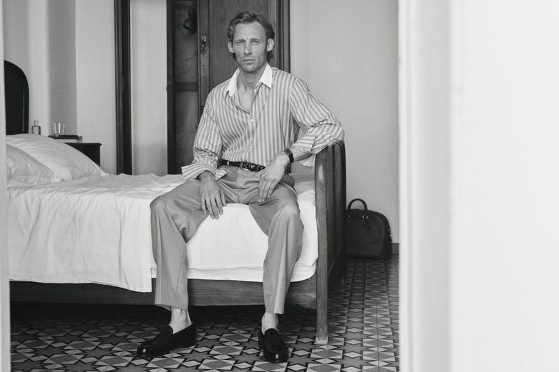 Black and white portrait of an adult male sitting on a bed in a collared, striped shirt and trousers, with a patterned floor and black bag.
