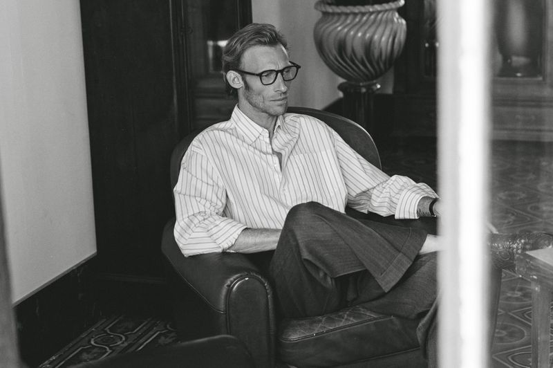 A man with glasses and a striped shirt sits in a leather armchair in a relaxed pose, captured in black and white with natural lighting.