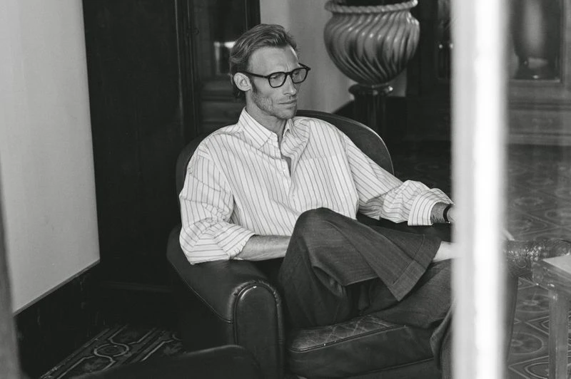 A man with glasses and a striped shirt sits in a leather armchair in a relaxed pose, captured in black and white with natural lighting.