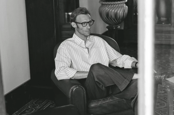A man with glasses and a striped shirt sits in a leather armchair in a relaxed pose, captured in black and white with natural lighting.