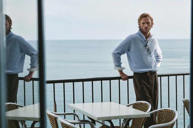 Adult male standing on a terrace overlooking the ocean wearing a blue button-down knit shirt and brown trousers with a belt and glasses.