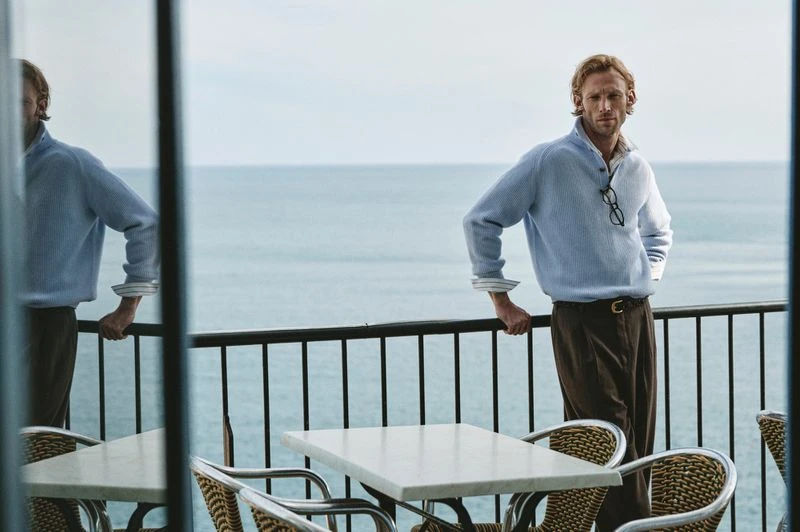 Adult male standing on a terrace overlooking the ocean wearing a blue button-down knit shirt and brown trousers with a belt and glasses.